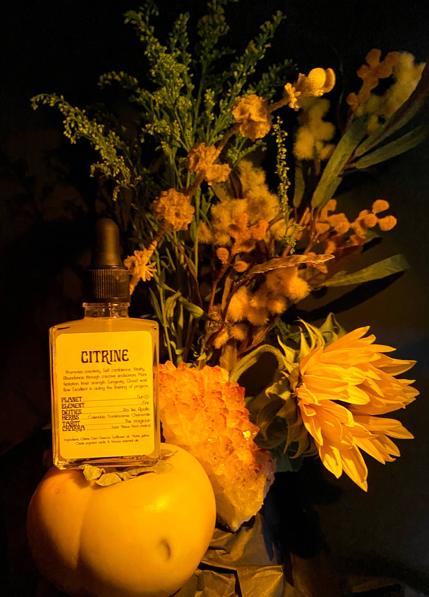 A clear glass bottle with a black dropper filled with yellow-colored gem essence oil, labeled 'Citrine', surrounded by yellow flowers, crystals, and yellow tomato