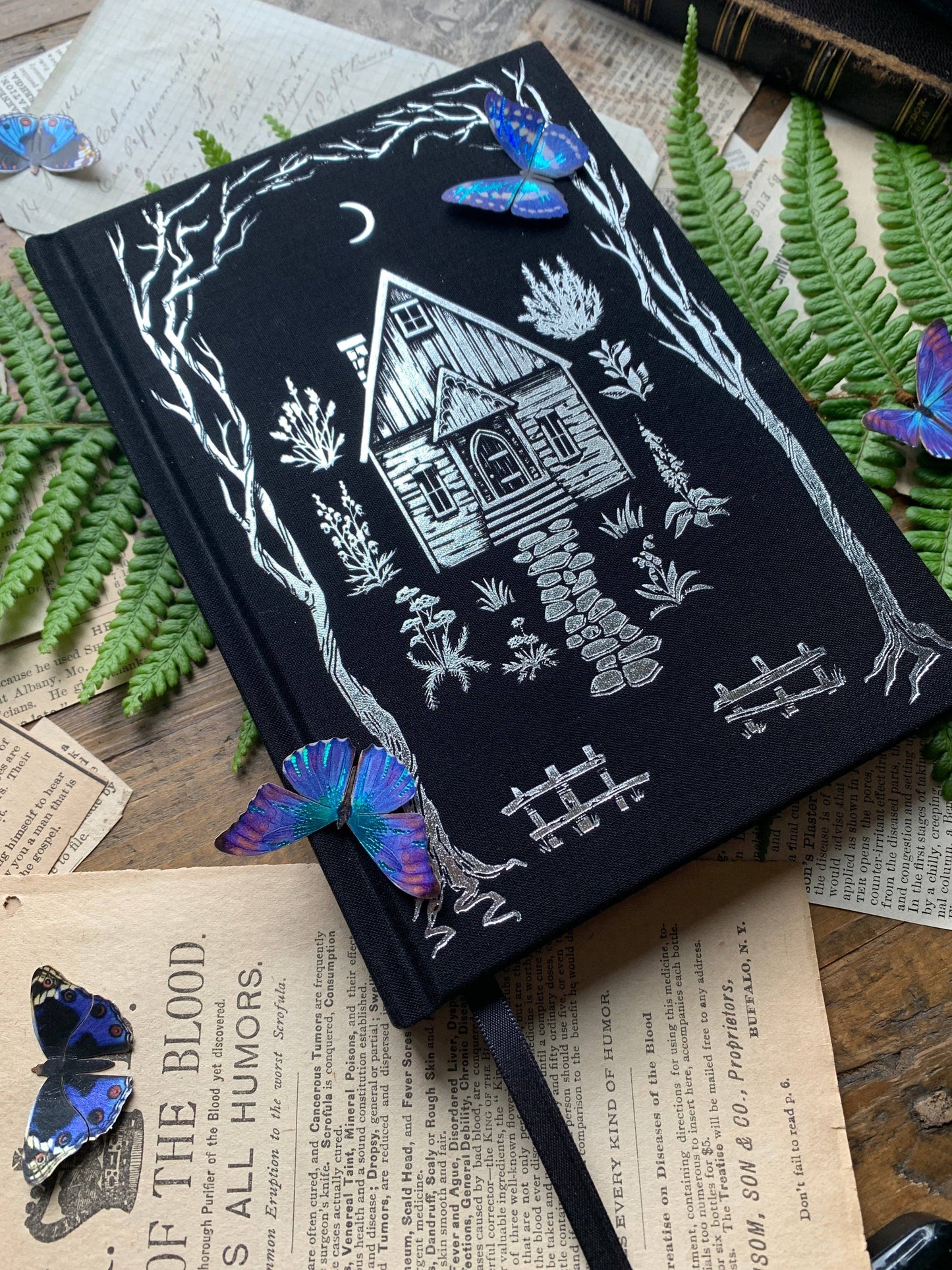 A black B6 vegan watercolor sketchbook with foil stamping, displayed on a wooden surface surrounded by butterfly decorations, fern leaves, and vintage paper.
