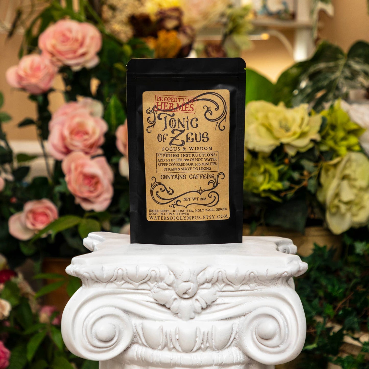 Tea package labeled 'Ionic of Zeus' on a decorative white pedestal with flowers in the background