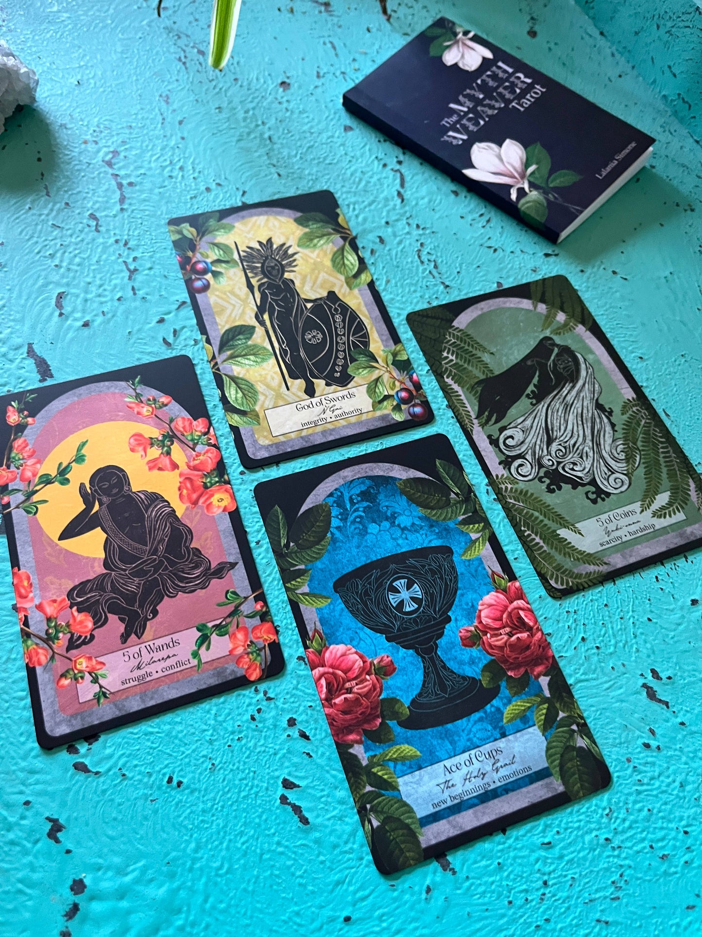 Four tarot cards on a blue textured surface with a book titled 'The Myth Weaver Tarot' in the background.