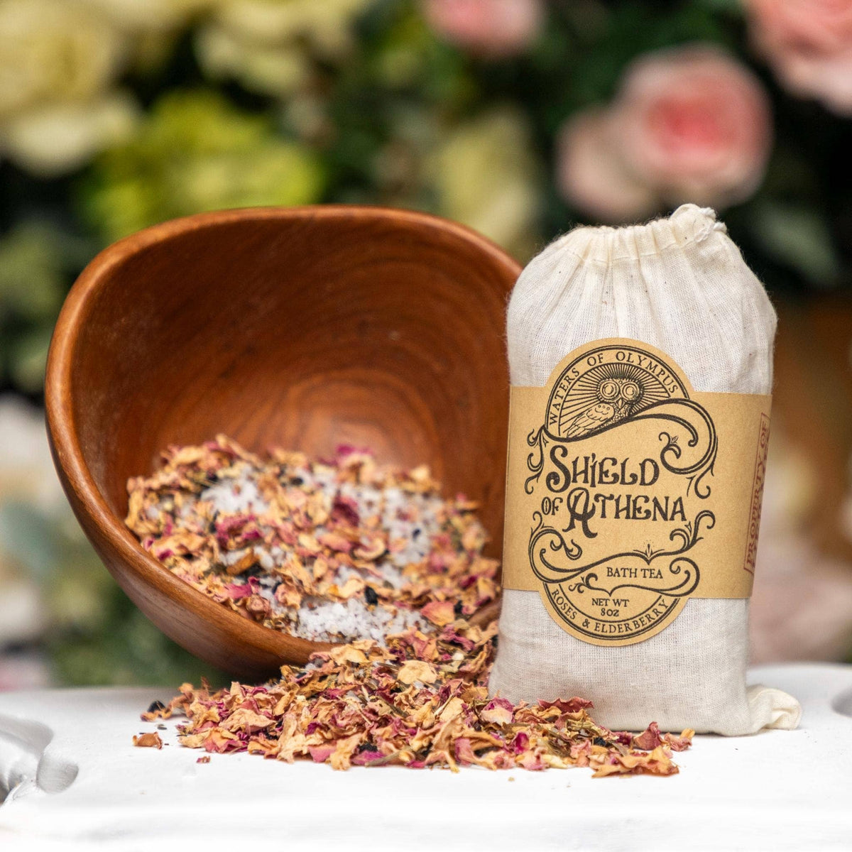 Bag of 'Shield of Athena' bath tea with a wooden bowl of bath salts on a blurred floral background