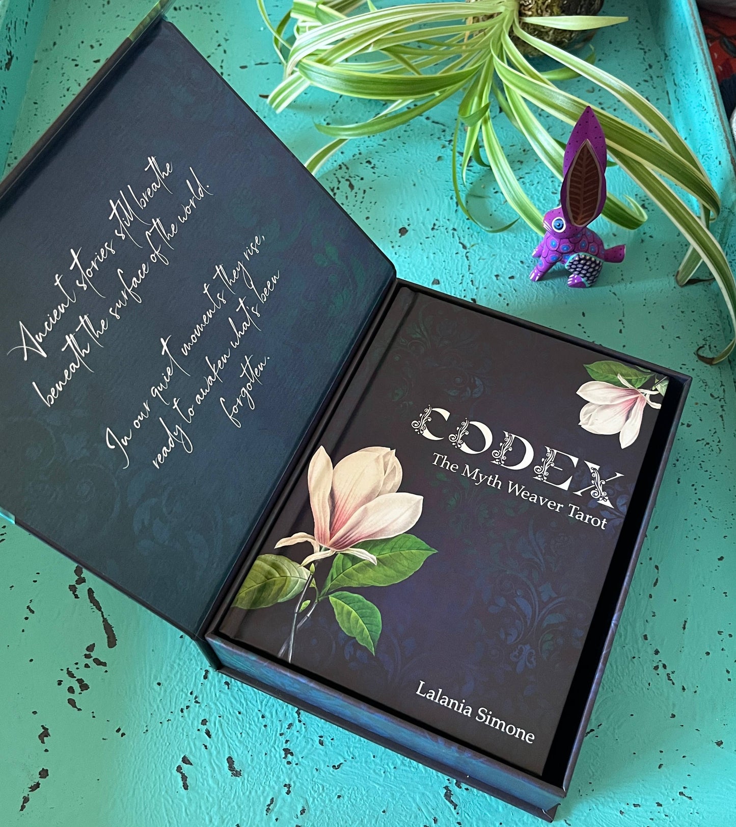 Open box of 'Codex: The Myth Weaver Tarot' by Lalania Simone on a teal surface with a plant and small figurine.