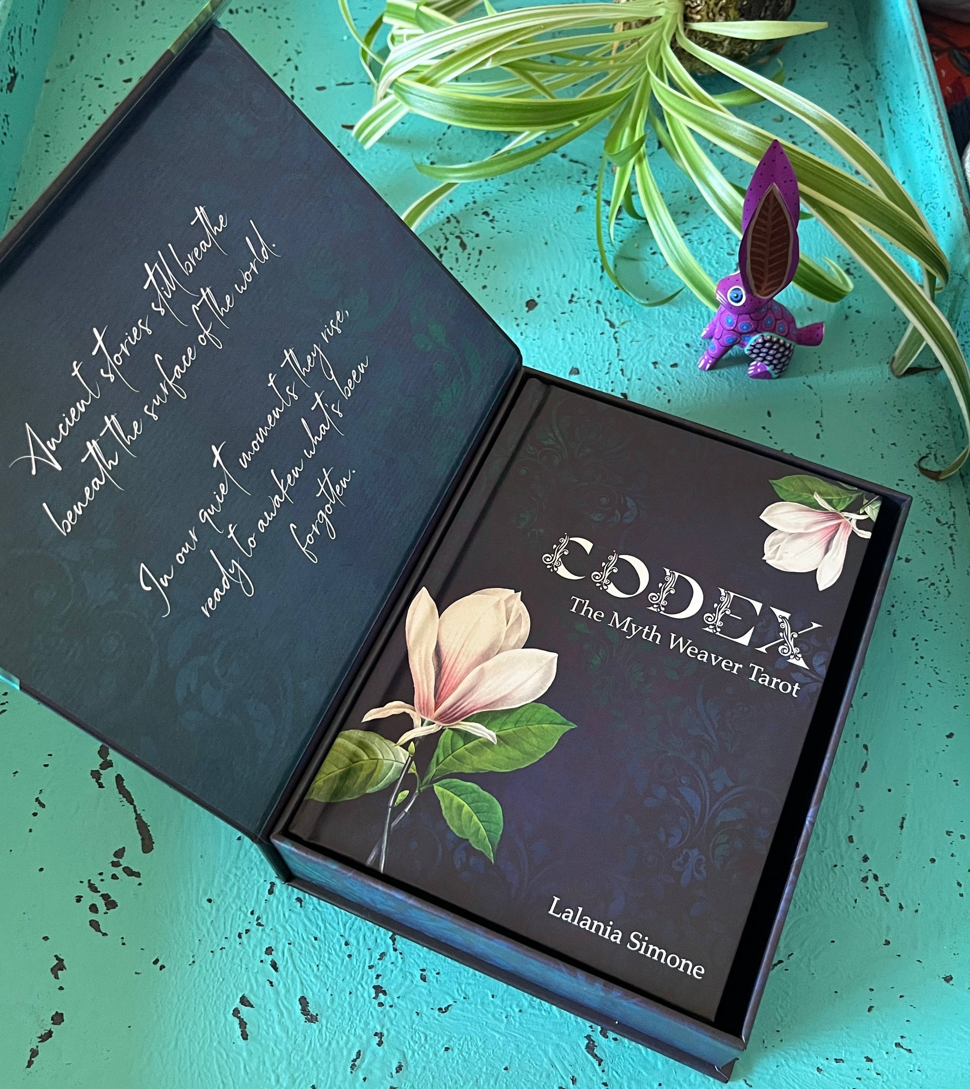 Open box of 'Codex: The Myth Weaver Tarot' by Lalania Simone on a teal surface with a plant and small figurine.