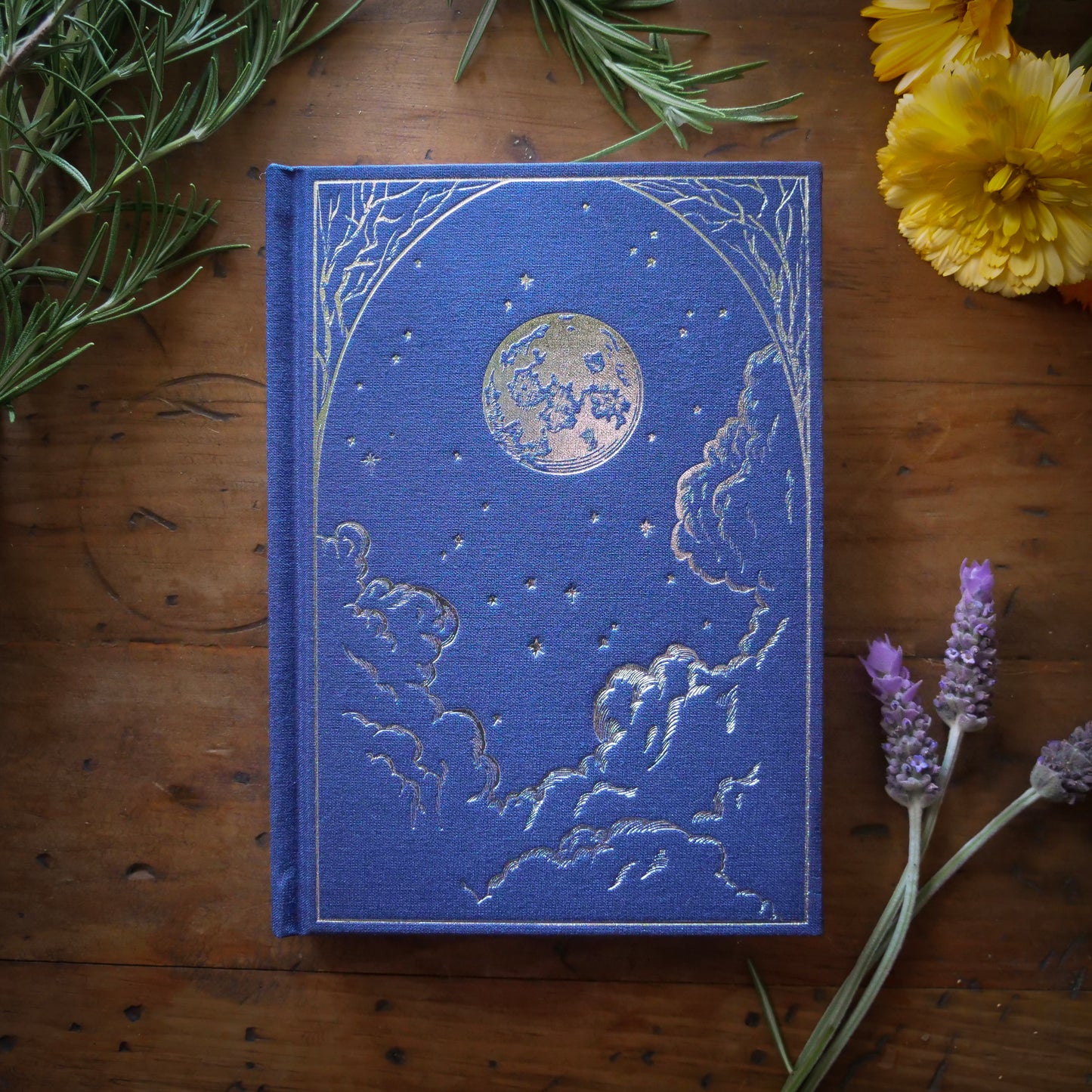 A dark blue B6 vegan watercolor sketchbook with foil stamping, displayed on a wooden surface surrounded by flowers and foliage