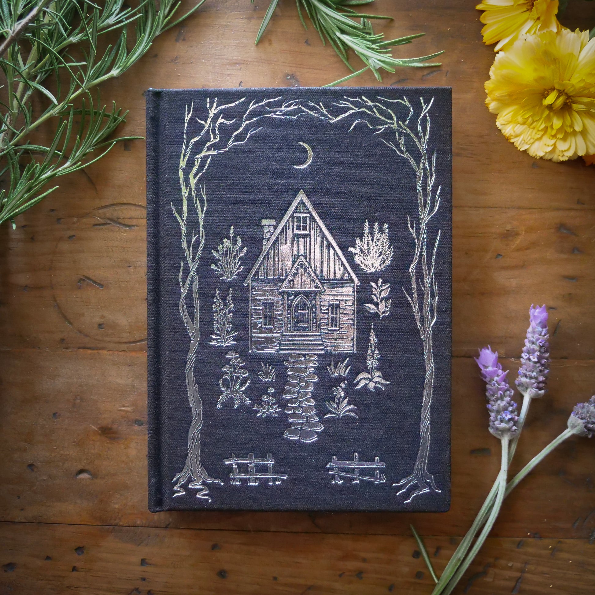A black B6 vegan watercolor sketchbook with foil stamping, displayed on a wooden surface surrounded by flowers and foliage
