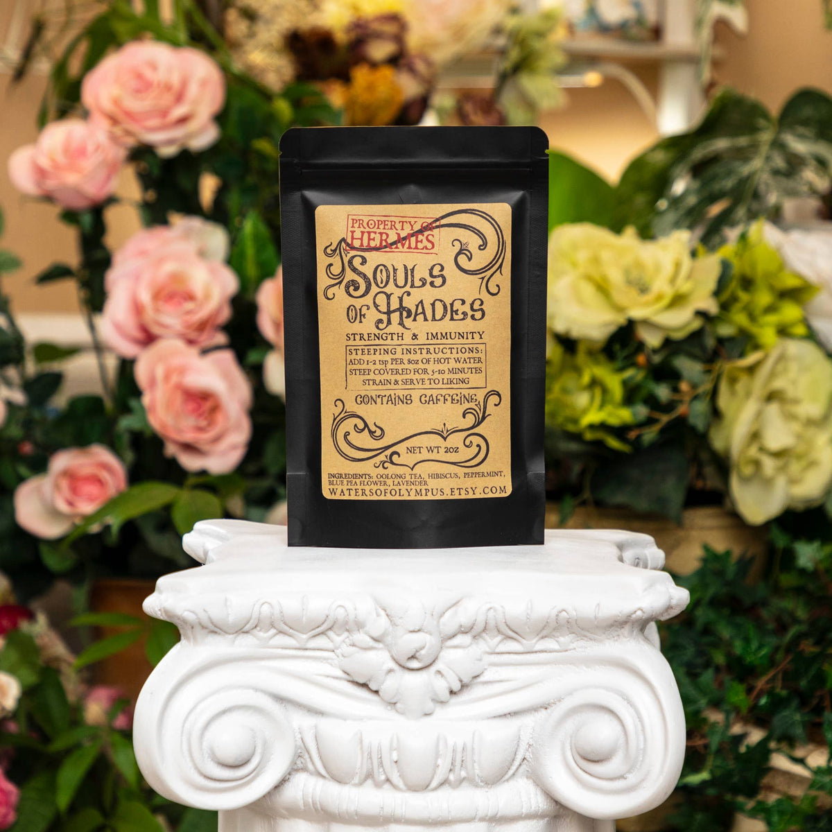 Black package labeled 'Souls of Hades' on a decorative white pedestal with flowers in the background