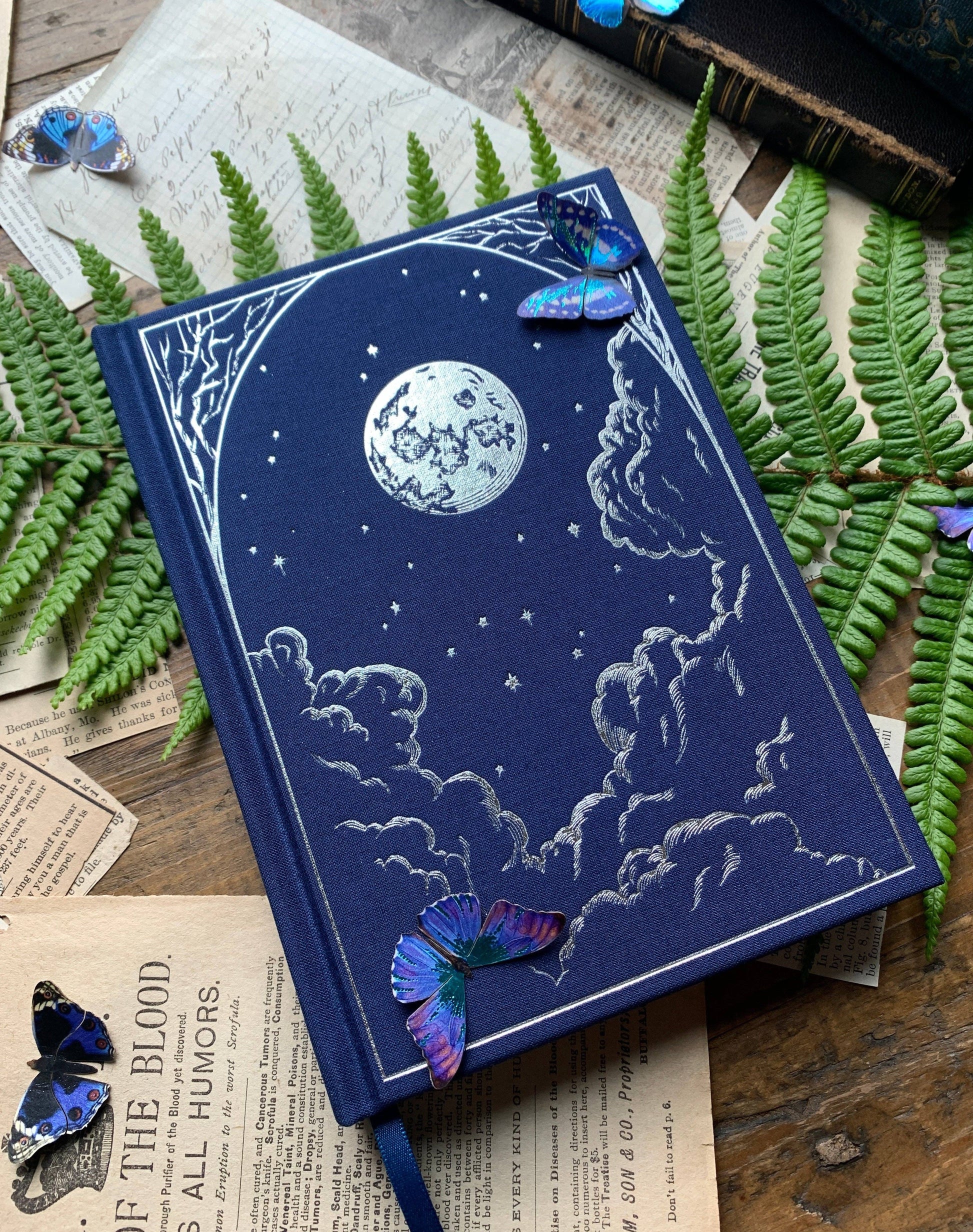 A dark blue B6 vegan watercolor sketchbook with foil stamping, displayed on a wooden surface surrounded by butterfly decorations, fern leaves, and vintage paper.